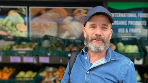 A man with a blue shirt and hat speaking to the camera with an array of fruit and veg behind him in a soft focus