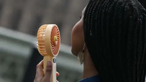 A young woman holds a battery-operated fan close to her face. 