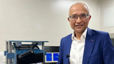 Dr Arnab Basu is wearing a blue suit and white shirt. He is smiling at the camera and wearing glasses. He standing next to the protoype machine that he has developed at his company Kromek.