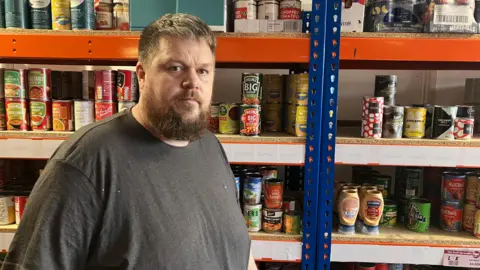 James Grant/BBC A man in a grey T-shirt stands in front of a three-storey shelf full of tinned food cans. 