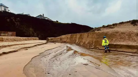 Newquay Coastguard Search and Rescue Team The picture shows a stretch of beach where heavy rainfall or recent flooding has carved a deep channel through the sand. The water has cut a sharp, steep-sided trench that runs from the foreground towards the back of the scene, leaving exposed sandy walls on either side. A person in high‑visibility waterproof gear stands to the right of the trench, looking down at the flowing water and the eroded sand. 
