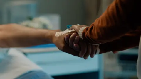 Getty Images Photo of two people holding hands in a hospital. 