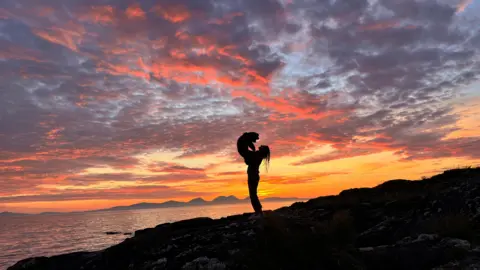 Pauline McAllister Vivid sky showing blue, orange, red and yellow at sunset with the shadowed outline of a woman holding a dog in the air, standing by the sea