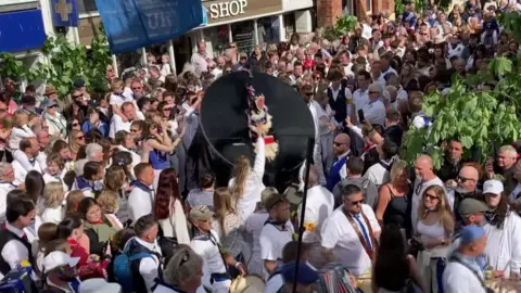 A crowd of people cheer and sing as a large black Obby Oss with blue ribbons dances through the streets of Padstow.