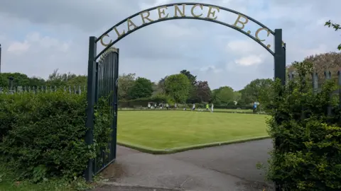 A metal arch with gold signage saying "Clarence B.C." above a bowling green, where people are playing bowls. There are trees surrounding the bowling green and hedges on either side of the arch.