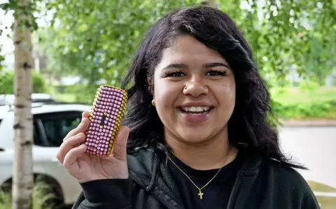 BBC News/Kristian Johnson Grace, wearing a black top and jumper, smiles as she holds up her brick-phone, which she has decorated in pink and yellow plastic gems