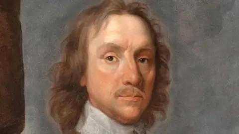 A 17th Century portrait of Oliver Cromwell, with shoulder-length curling mid-brown hair and moustache, wearing a white lace collar done up at the neck over a yellow under-jacket, above which is grey armour. He looks at the viewer with dark eyes.