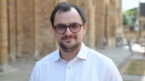 Joe Bilton/BBC A head and shoulders shot of a man with a short moustache and beard, short brown hair, brown circular rimmed glasses and a white shirt. The background is blurred but it is the wall of a cathedral.