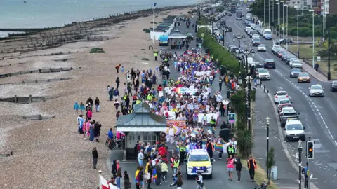 An aerial image of a Pride procession moving along a seafront - a pebble beach is on the left with a road to the right