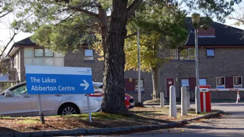 A sign indicates the direction of The Lakes and Abberton Centre at a hospital. Bollards and a tree are next to the sign.