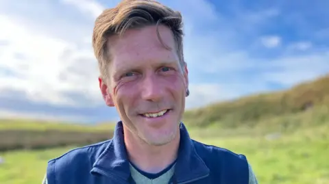 Ulf Keller looking to the camera on a sunny day on Rathlin Island.  He has short dark-blond hair and is wearing a blue bodywarmer over a green jumper and navy T-shirt. 