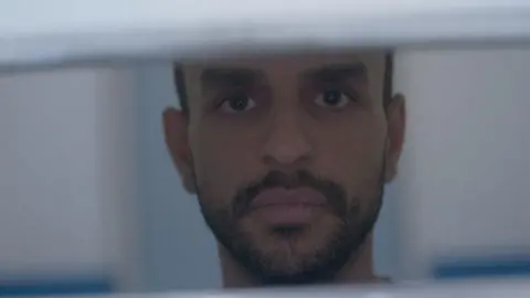 A close up of Mohammed Altaher's face as he looks through bars. He has short black hair and a close-cropped black beard. 