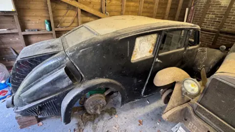 A 1934 Crossley Streamline car in a barn. It is black and covered in dust, with its bonnet facing a brick and wood wall. It is resting on bricks. On the right is the front of another vintage car. 