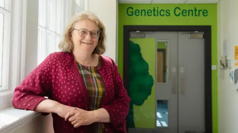 Friends of ANCHOR Prof Zosia Miedzybrodzka is standing in a brightly lit corridor, in front of a green door with the words 'Genetics Centre' above. She has light brown hair and is wearing glasses, a tartan top and a red cardigan. She is smiling at the camera