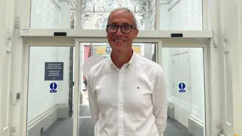 Simon Youden is standing in front of a glass door entrance. He is wearing a white button up shirt. He is holding his arms behind his back.