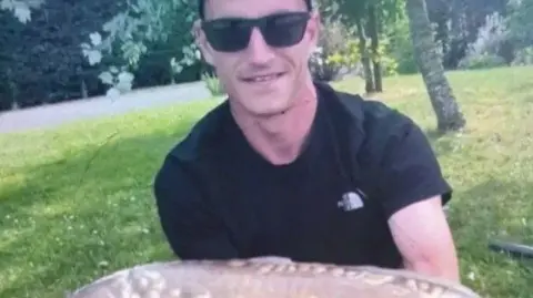 Louis Price is wearing a black T-shirt and dark sunglasses. He is holding a large fish close to the camera and smiling. There is greenery and trees in the background.