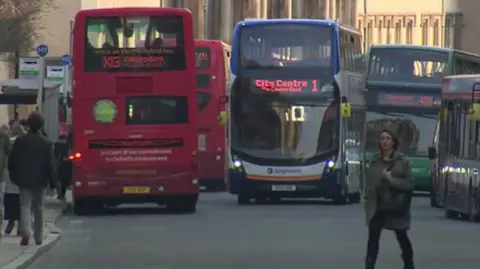 A busy Queen Street in Oxford with many buses on the road and people crossing the street and walking on the pavements. A bus is coming towards the camera with a sign reading "City Centre". Another one is travelling away, with a sign reading "Abingdon".