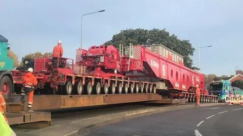 Suffolk County Council A general view of an abnormal load on a road. Highways workers in orange hi-vis  coats and trousers stand near or on the abnormal load. 