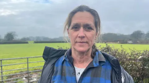 A picture of a lady with brown hair. She is wearing a blue chequered shirt and a black jacket.