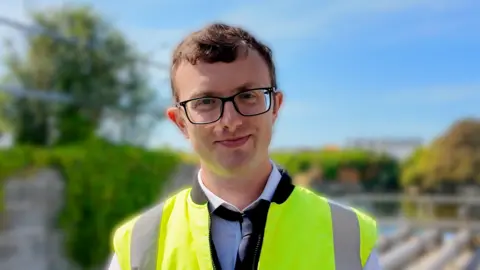 Daffyd Griffiths pictured wearing a white shirt with a black tie and a high viz yellow jacket with no sleeves. He has short brown hair and square black glasses. 