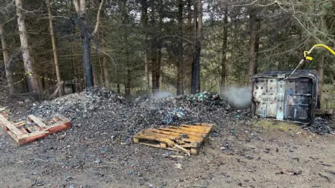 Nottinghamshire Fire and Rescue Service A large pile of scorched lithium-ion batteries that had been discarded near woodland. Two wooden pallets are also amongst the waste pile and a metal crate on its side is on the right of the frame, with a thin yellow tube misting down the scene.