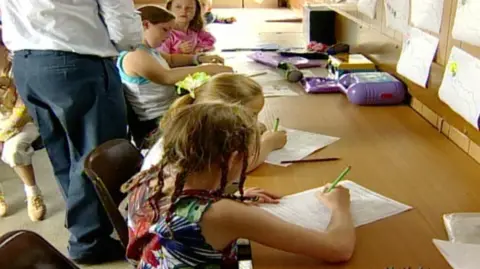 A still from a BBC TV report. Several children are writing as they sit along a wooden desk. A man, likely the teacher, is standing next to them.