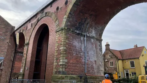 A person wearing a reflective jacket looks up at the viaduct, built of red brick and has arches. It is cordoned off as repairs continue.