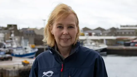 Head and shoulders of Elspeth Macdonald. She has blond hair and is wearing a blue jacket with a stylised drawing of a fish on the jacket under the right shoulder. She is standing in a small harbour which is visible in the background with a few small fishing boats there.