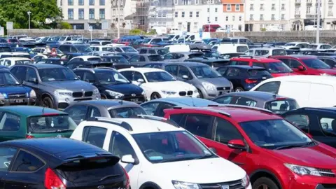 Cars fill all the spaces at a car park with a wall and harbour buildings beyond.