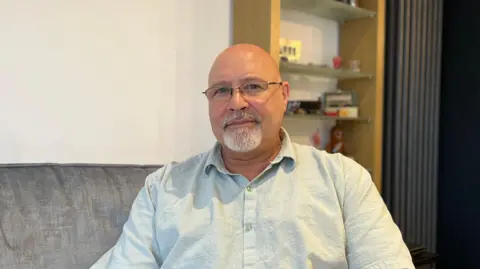 A man with a shaved head, glasses and a grey goatee-style beard. He is wearing a light linen shirt with the top-button unbuttoned. He is sitting on a dark grey sofa, in front of a wooden shelving unit. He is staring directly at the camera.