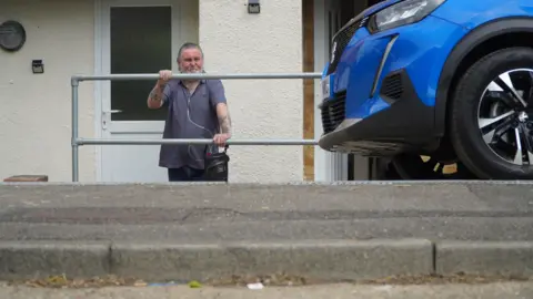 Michael Partridge, wearing a blue shirt and black trousers, stands outside his house in Medway, leaning on a metal barrier and behind a steep kerb. His white front door is behind him and his blue car is parked on his left.