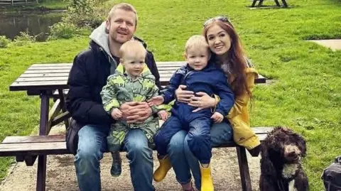 A family of four sit on a picnic bench, with a dog.