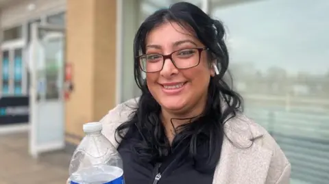 A woman with long black hair and wearing glasses and a grey coat is on a paved area smiling at the camera. She is holding a bottle of water and a building in the background is blurred.