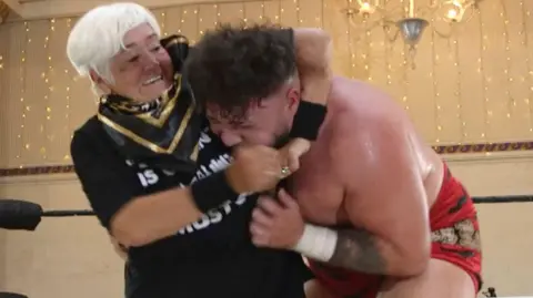 Susie Matthews, 73, taking part in wrestling showcase in Cardiff. She has a male professional wrestler in a headlock.