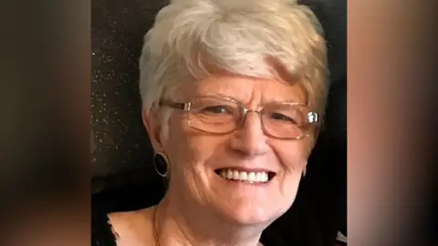 Joan Gansh with short light hair and glasses, wearing a leopard print top and a necklace.