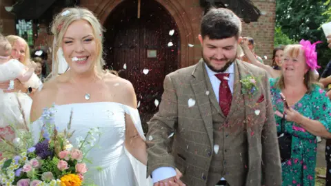 Kym and Tyler A married couple are pictured smiling, leaving a church, and friends and family are throwing confetti over them. The bride, on the left, is wearing a white dress and carrying flowers. The groom is wearing a moss green checked suit with waistcoat.
