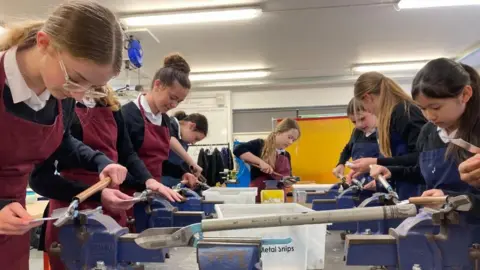 Eight students at Hayesfield Girls' School in Bath are at a table affixed with vices, using them to cut metal in to shapes. 