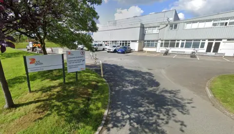 The image shows an orange and white sign which says DS Smith, behind which can be seen a white and grey industrial building with a tree and grass to the left.
