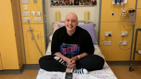 Isabel Marsh Isabel Marsh sat on a hospital bed. She is smiling at the camera and is wearing a black top and leggings. 