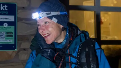 Anna Troup at the finish line at Kirk Yetholm. She is in front of a building and she is looking to the side and smiling. She is wearing a navy woolly hat and a headtorch, which is lit. She also wears a bright blue outdoor jacket.