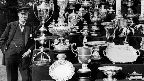 Bettmann/Getty A man dressed in a nautical cap standing next to a large collection of trophies