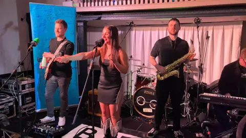 A band performs on a small BBC stage. A woman wearing a black dress is standing in the middle and smiling while pointing. Two men are either side of her, one with a guitar and another with a saxophone. A man on a keyboard can be seen on the right.