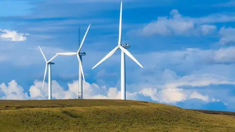 Scout Moor wind farm showing three large white wind turbines on a green grassy area with blue sky and white clouds in the background