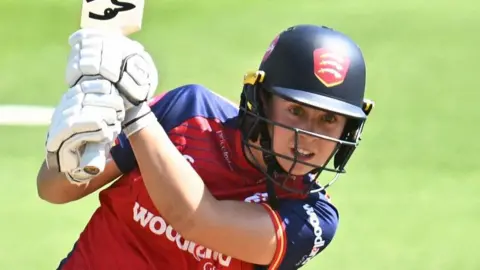 Jodi Grewcock wearing an Essex Cricket jersey and helmet swings her bat during a match.