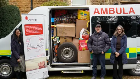 An ambulance with its side door is open. Lots of cardboard boxes and plastic bags are stacked on the inside and a spare tyre can be seen. Three people, a man and two women stand next to it. 