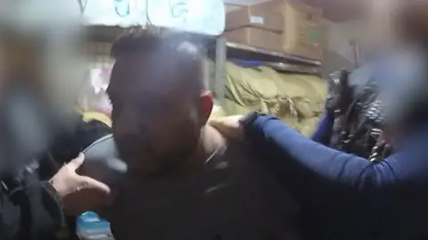 GMP Rawal Rehman, wearing a grey long-sleeved top, is held up by two police officers with his arms handcuffed behind his back. 