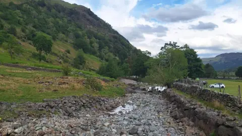 West Cumbria Rivers Trust A section of the Upper Derwent. Much of the riverbed is dry with stones visible along a large stretch. There are fields to either side along with hills and trees. The sky is blue but with some areas of cloud.