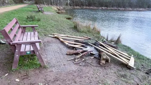 Woolsington Parish Council The remains of a fire next to a bench facing a pond. Uprooted fence posts and tree logs have been thrown on top of the fire.