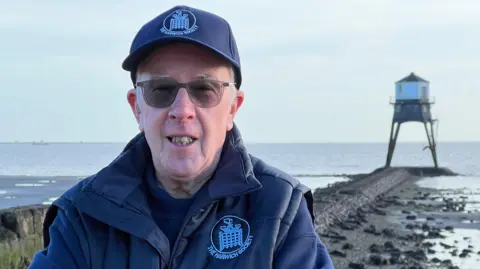 Andy Schooler is wearing glasses and a navy cap, jumper and gilet. Behind him, a rocky causeway leads to one of the lighthouses, which is mostly surrounded by water has a white building on top of iron legs.
