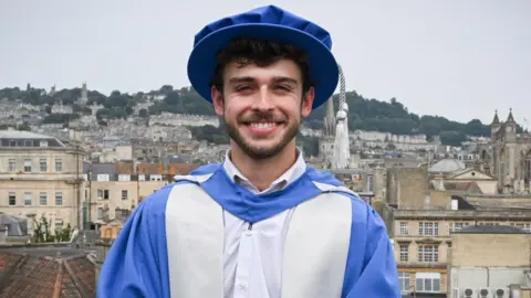 George Clarke is standing on a balcony overlooking the city of Bath. He is dressed in a graduation cap and gown after being given an Honorary Doctorate of Arts. The gown and cap are blue and silver. George has short dark hair and facial hair and is standing with his hands clasped in front of him, looking at the camera and smiling.